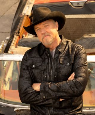 Trace Adkins wearing a black leather jacket and cowboy hat, standing with arms crossed in front of an old car.